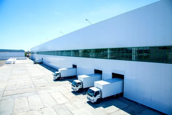 Modern warehouse with three white delivery trucks parked at loading docks, glass windows, and a sunlit concrete service yard.