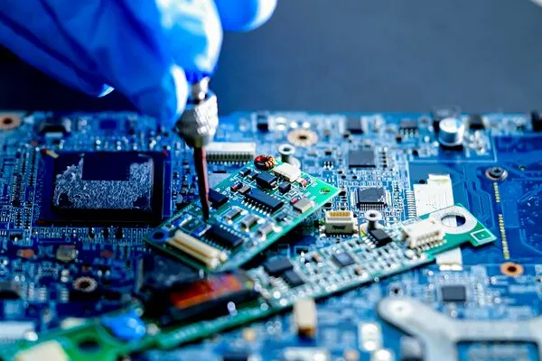 Gloved technician uses a screwdriver to repair a small circuit board on a blue motherboard, highlighting precision electronics work.
