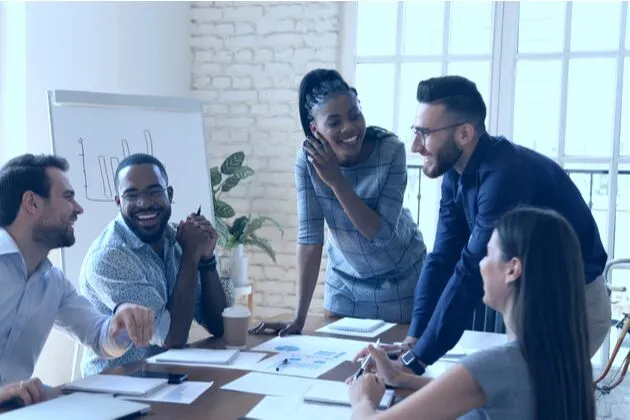 Diverse team of five coworkers smiling and collaborating around a meeting table in a bright office, with documents, coffee, and a whiteboard.